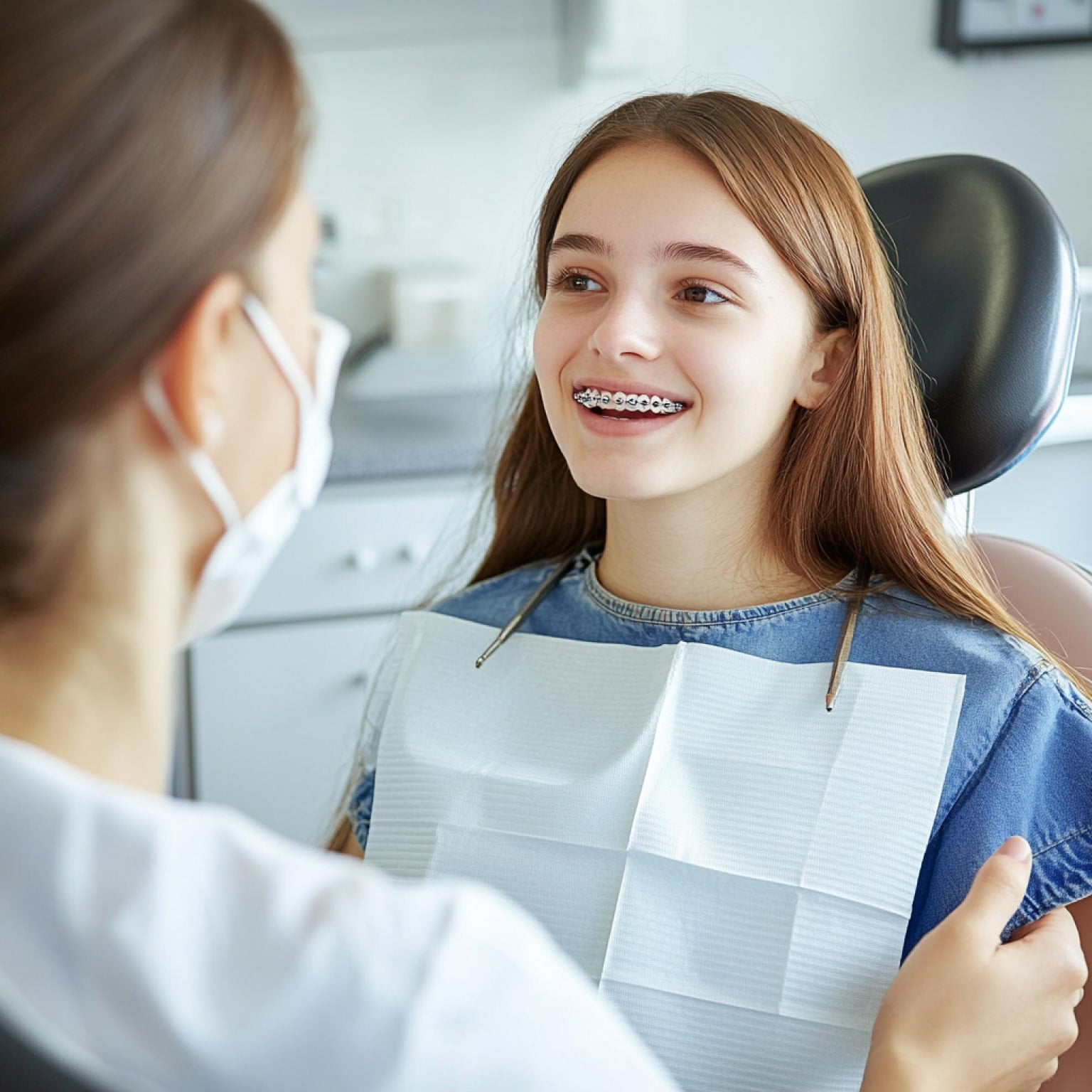 A person with long hair, wearing braces, sits smiling in a dental chair, interacting with a dental professional in a clinic.
