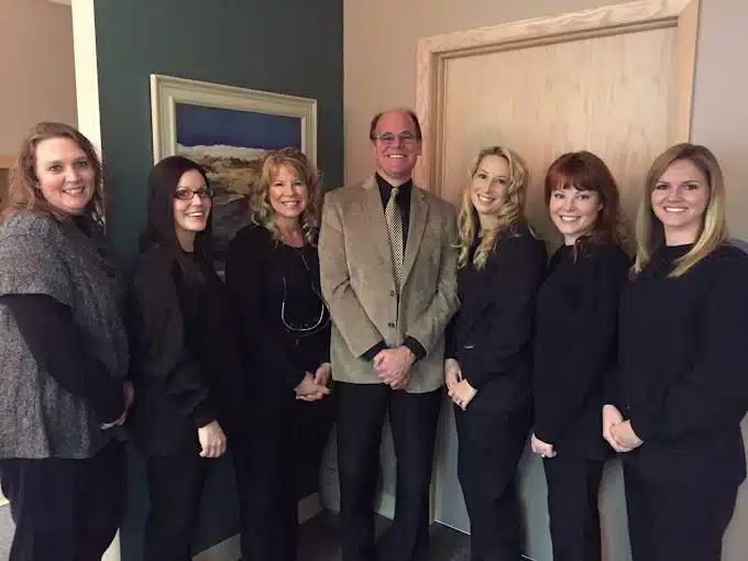Seven people smiling indoors, standing in a line. One person wears a tan blazer, others wear dark attire. A landscape painting hangs on the wall.