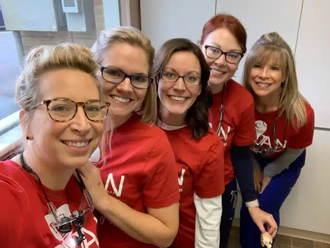 Five smiling people in red shirts pose for a group photo indoors, with a window in the background and white cabinets behind them.