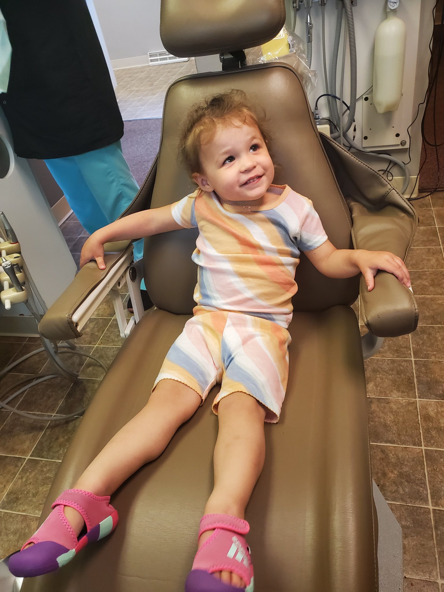 A child in colorful attire sits smiling on a dentist chair, with a person in scrubs partially visible in the background.