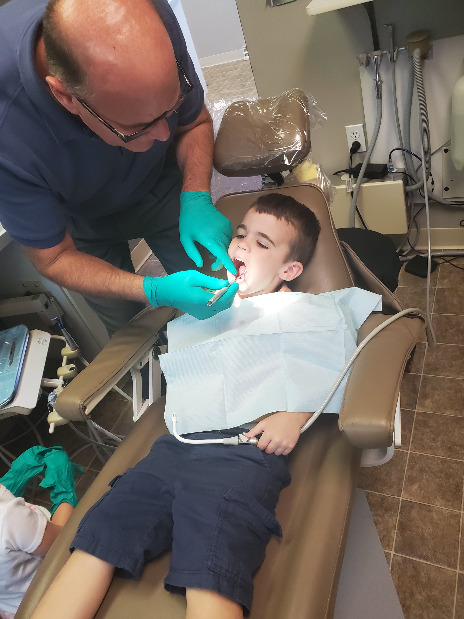 A person in a dental office examines a child's mouth, while the child lies on a reclined chair. Dental equipment surrounds them.